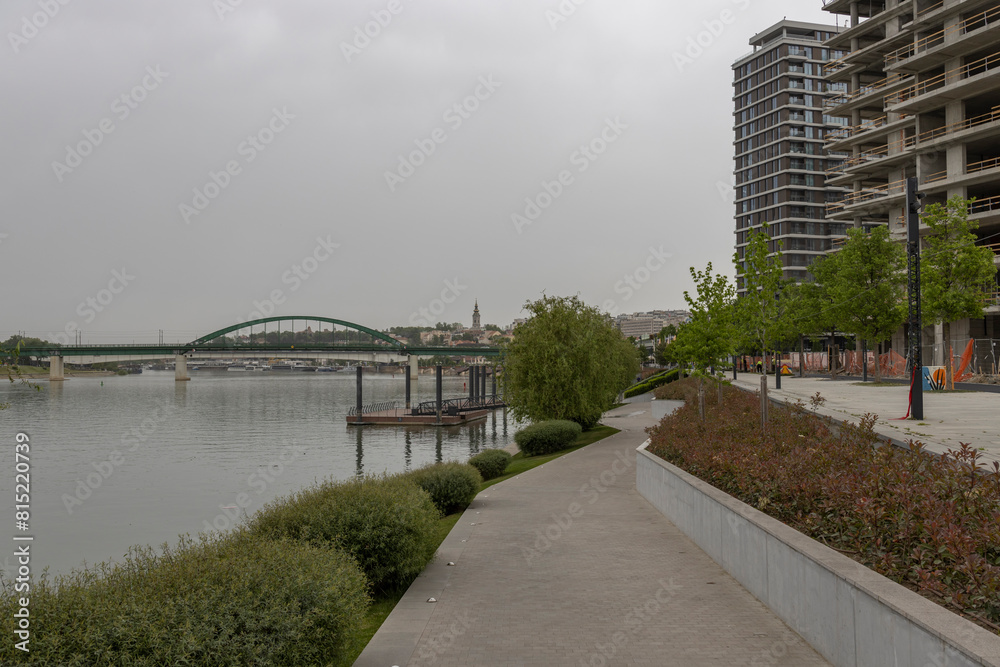 Fototapeta premium A concrete walkway along a riverfront with high-rise buildings in the background, featuring a bridge and greenery, under an overcast sky.