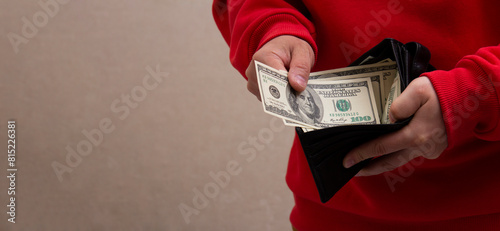 Man in red hoodie pulls out 100 USD. Dollar bill from black leather wallet. Horizontal banner with copyspace for text. Concept of travel, tourism, debt payment, mortgage, credit. Cash money of USA
