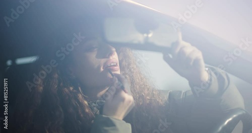 A young woman wearing braces is captured applying lipstick in a car, using the rearview mirror for assistance, reflecting a moment of everyday beauty rituals. photo