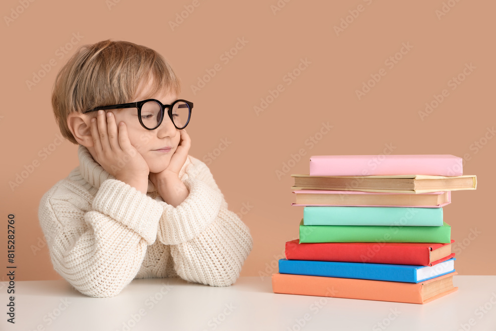 Cute little boy in eyeglasses with stack of book sitting at table against color wall Cute little boy in eyeglasses with stack of book sitting at table against color wall