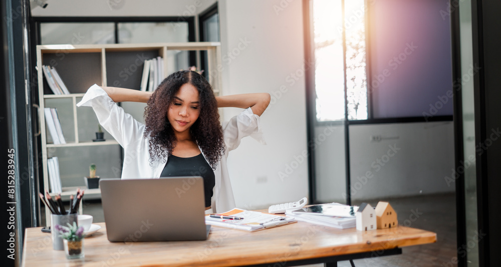 A woman is sitting at a desk with a laptop and a stack of papers. She is wearing a white shirt and black tank top. The room is well lit and has a modern design. The woman is focused on her work A woman is sitting at a desk with a laptop and a stack of papers. She is wearing a white shirt and black tank top. The room is well lit and has a modern design. The woman is focused on her work