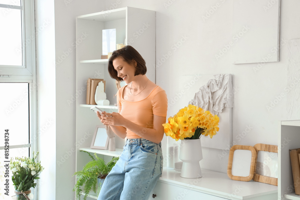 © Pixel-Shot - Young woman using mobile phone with daffodils in vase on commode at home