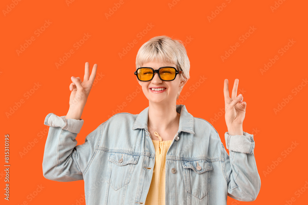 Portrait of beautiful young woman with sunglasses showing victory gestures on orange background