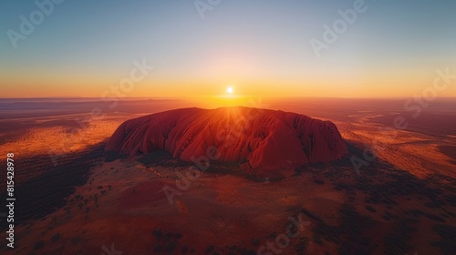 Aerial view of Uluru (Ayers Rock) in Australia, with its massive red sandstone formation rising from the flat desert landscape at sunset. 