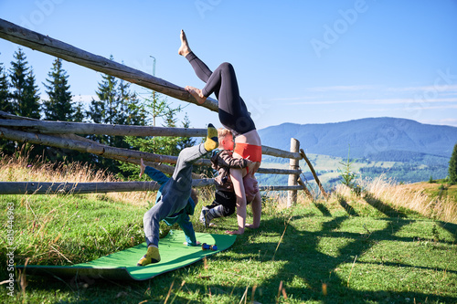 Mother and her children doing yoga outdoors in scenic mountainous landscape in the morning. Woman and boy inverted in headstand, near rustic wooden fence in background.