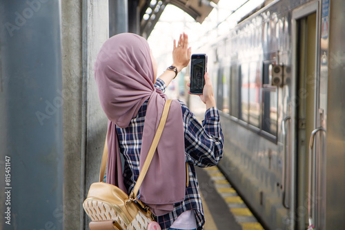 back view of asian muslim woman holding smartphone waving goodbye at train station platform. traveling concept