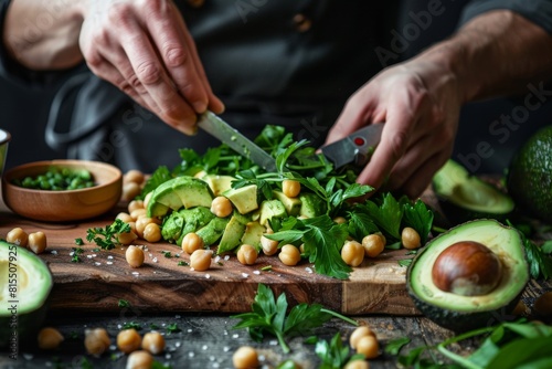 A person's hands carefully preparing plantbased food, such as avocado and chickpeas on wooden board, surrounded by fresh vegetables in dark kitchen background. photo