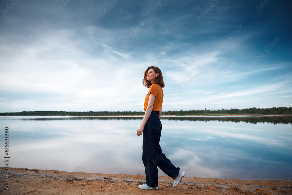 Red-haired girl in an orange T-shirt and orange glasses on the shore of a lake Red-haired girl in an orange T-shirt and orange glasses on the shore of a lake