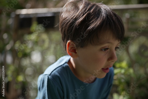 A thoughtful young boy, wearing a blue shirt, is shown in a natural outdoor setting. His expression is pensive as he looks down, with the sunlight highlighting his hair and creating 
