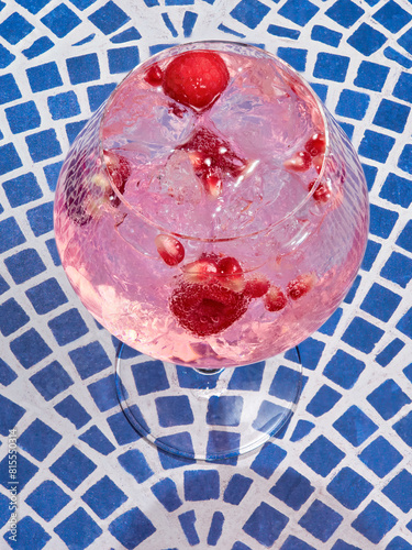 A close-up of a refreshing pink gin tonic filled with ice and berries set against a blue tile background