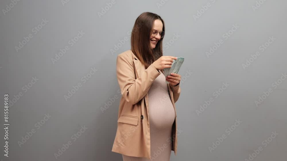 Rich happy pregnant woman wearing dress jacket and glasses isolated over gray background holding big sum of euro banknotes counting her salary Rich happy pregnant woman wearing dress jacket and glasses isolated over gray background holding big sum of euro banknotes counting her salary