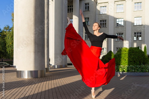 Dancing Flexible Winsome Professional Caucasian Ballet Dancer in Red Skirt in Leg Stretching Dance Pose With Lifted Hands Against Pillars