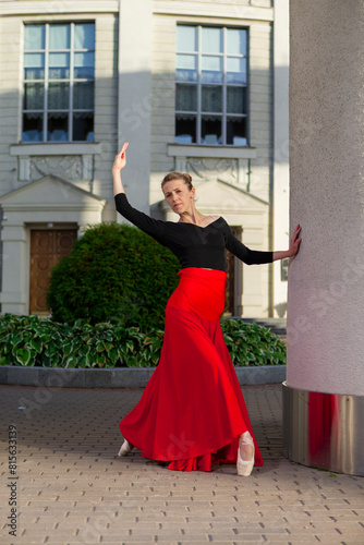 Tranquil Flexible Winsome Professional Caucasian Ballet Dancer in Red Skirt in Leg Stretching Dance Pose With Lifted Hand Against Pillars in City.