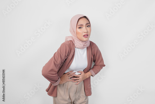 An unwell sick young Asian Muslim woman placing hand on stomach suffers pain isolated on white color background. © Reezky