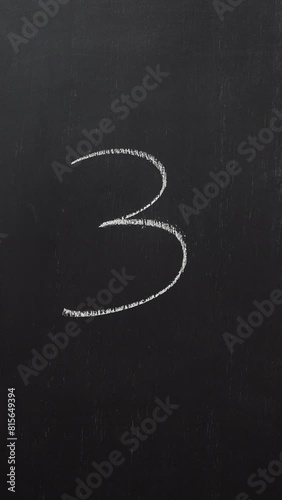 Hand writes the number 3 with white chalk on a black board. Vertical video