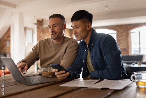 Gay couple sitting in kitchen using a laptop and smartphone for personal finances photo