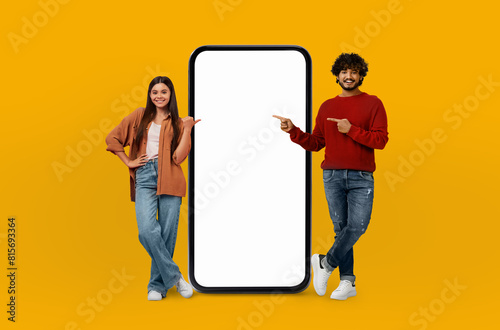 Multiethnic young lovers are standing next to a large phone, appearing to be engaged in conversation or possibly using the phone. The setting is nondescript but suggests a public space.