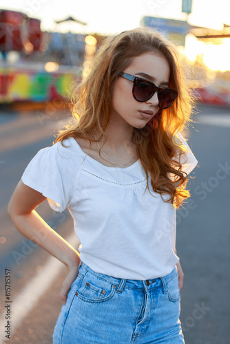 Fashionable beautiful hipper redhead girl in stylish casual clothes with a white T-shirt and jeans with a fashion frame of sunglasses walks in the city at sunset photo