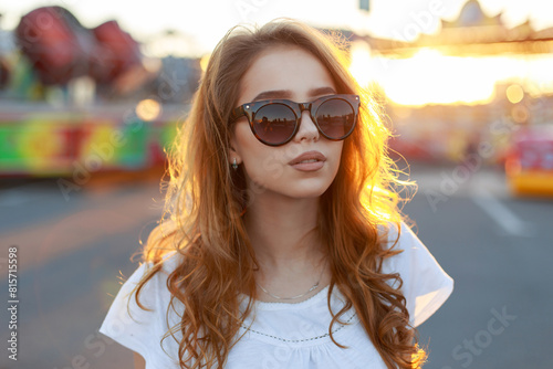 Beautiful fashionable female portrait of a hipster pretty girl with red hair and sunglasses in a fashionable suit at sunset