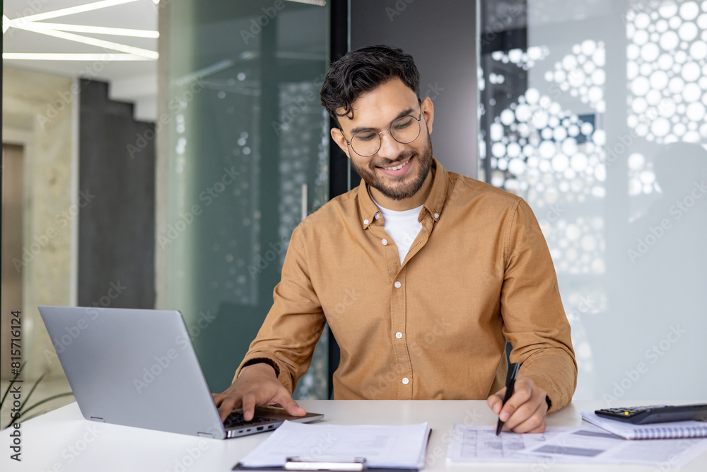 Obraz premium Smiling businessman working on laptop and taking notes in modern office