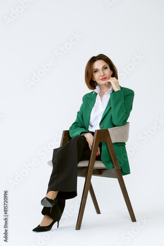 Attractive middle-aged business woman in black trousers and a green jacket is sitting on a chair on a white background. Portrait of a business lady.
