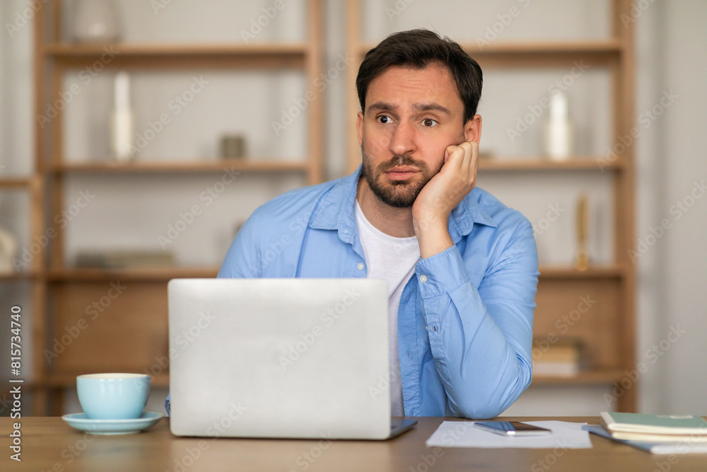 A young man is seated at a desk in a well-lit room, showing signs of boredom or fatigue as he rests his cheek on his hand while staring blankly at a laptop screen A young man is seated at a desk in a well-lit room, showing signs of boredom or fatigue as he rests his cheek on his hand while staring blankly at a laptop screen