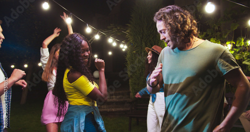 Happy beautiful mixed-race girls dancing in garden at party. Caucasian girl in hat and boy making funny moves. African American female dance. Happy guy moving to music. Celebration concept