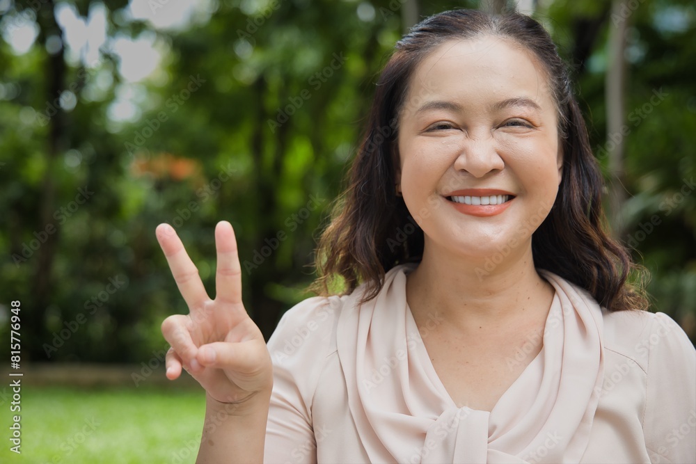 Happy smiling middle aged asian woman pointing 2 fingers up, concept of v for victory, two points, number two, second point in summer green park Happy smiling middle aged asian woman pointing 2 fingers up, concept of v for victory, two points, number two, second point in summer green park