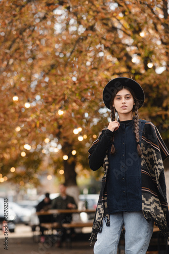 A fashionable girl showcases boho elegance, topped with a black hat, amidst the autumn city streets.