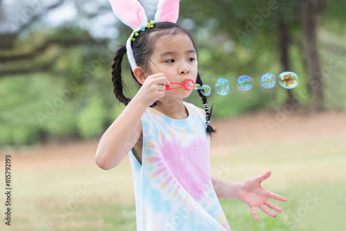 Happy 6 year old Asian little girl with bunny ears blowing soap bubbles in park, having fun, sunny day. Cute child playing outdoors in summer in nature