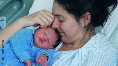 New 30s mother kissing newborn baby on forehead bonding with infant right after childbirth resting at hospital bed during the initial days of son's life, motherhood and maternal care concept