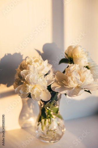 White peony flowers in glass vase in soft sunset light. Delicate beauty of white peonies bathed in the golden sunset casting soft shadows. A serene composition spring blooming. Trendy sun light.