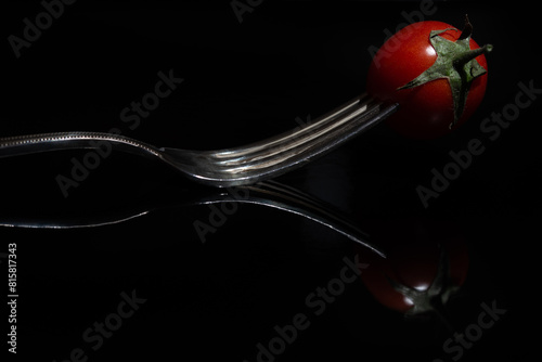 Close-up of a small red tomato stuck on a metal fork. The fork and the tomato are reflected against a dark background