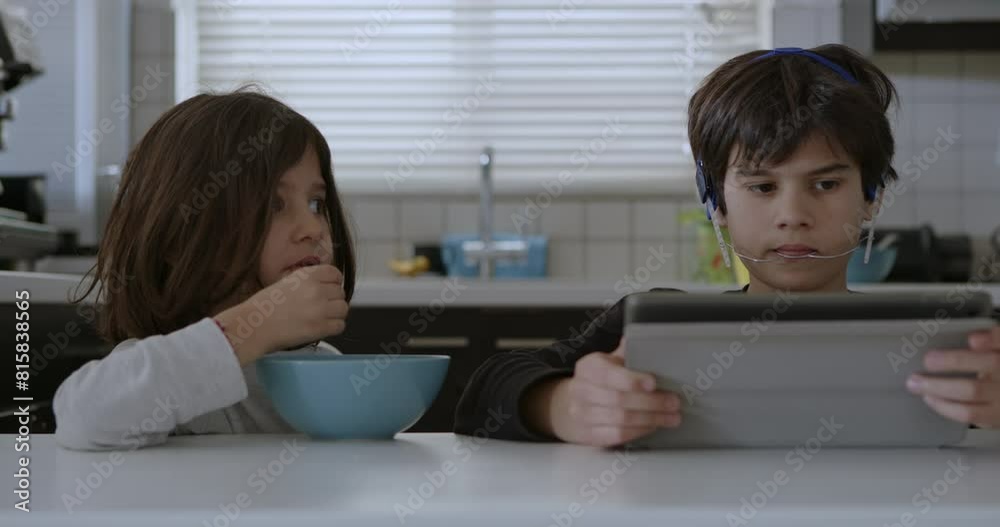 A boy with mouth braces is engrossed in his digital tablet while sitting at the kitchen table, as another child eats breakfast beside him. A boy with mouth braces is engrossed in his digital tablet while sitting at the kitchen table, as another child eats breakfast beside him.