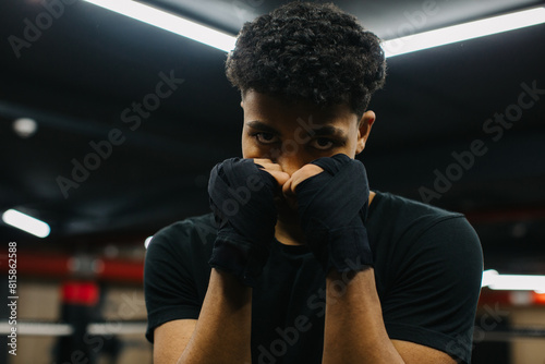 A determined young boxer is looking at the camera while standing in a fighting stance with boxing bandages on his fists. Sporty young man training in boxing gym.