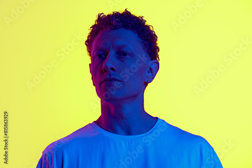 Portrait of man with curly hair standing with serious face, looking, posing against yellow background with blue neon light. Concept of human emotions, facial expression, lifestyle, casual fashion