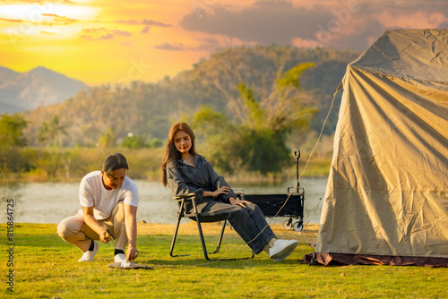 Young asian couple preparing tent outdoor in forest near lake while Beautiful woman relax in camping chairs under dramatic evening sky. Colorful sunset in mountains. Camping traveler concept. 