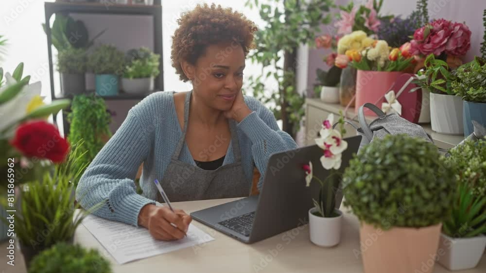 A smiling black woman taking notes in a colorful indoor flower shop, surrounded by plants and a laptop. A smiling black woman taking notes in a colorful indoor flower shop, surrounded by plants and a laptop.