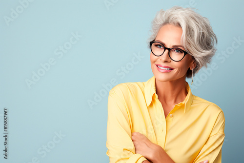 Portrait of a beautiful business elderly woman with short gray hair wearing glasses and a yellow blouse on a blue background, copy space