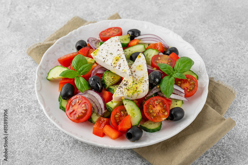 Greek salad of fresh juicy vegetables, feta cheese, herbs and olives on a white plate on a light concrete background. Healthy food. Copy space, top view.
