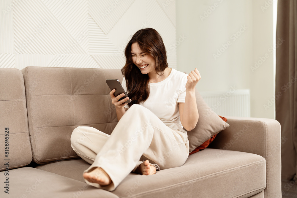 Woman Celebrating Success On Smartphone In Living Room: Happy, Excited, Home Interior, Casual Lifestyle Woman Celebrating Success On Smartphone In Living Room: Happy, Excited, Home Interior, Casual Lifestyle