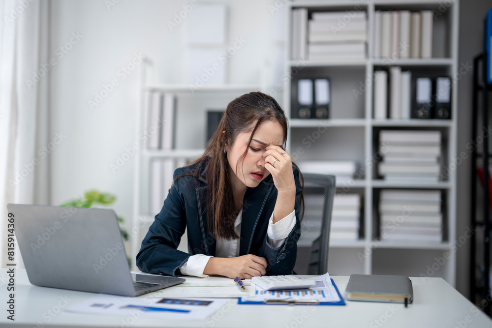 A woman is sitting at a desk with a laptop and a stack of papers. She is looking at the papers and she is in a state of distress A woman is sitting at a desk with a laptop and a stack of papers. She is looking at the papers and she is in a state of distress