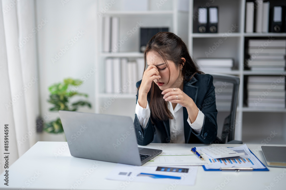 A woman is sitting at a desk with a laptop and a stack of papers. She is looking at the laptop and she is in a state of distress A woman is sitting at a desk with a laptop and a stack of papers. She is looking at the laptop and she is in a state of distress