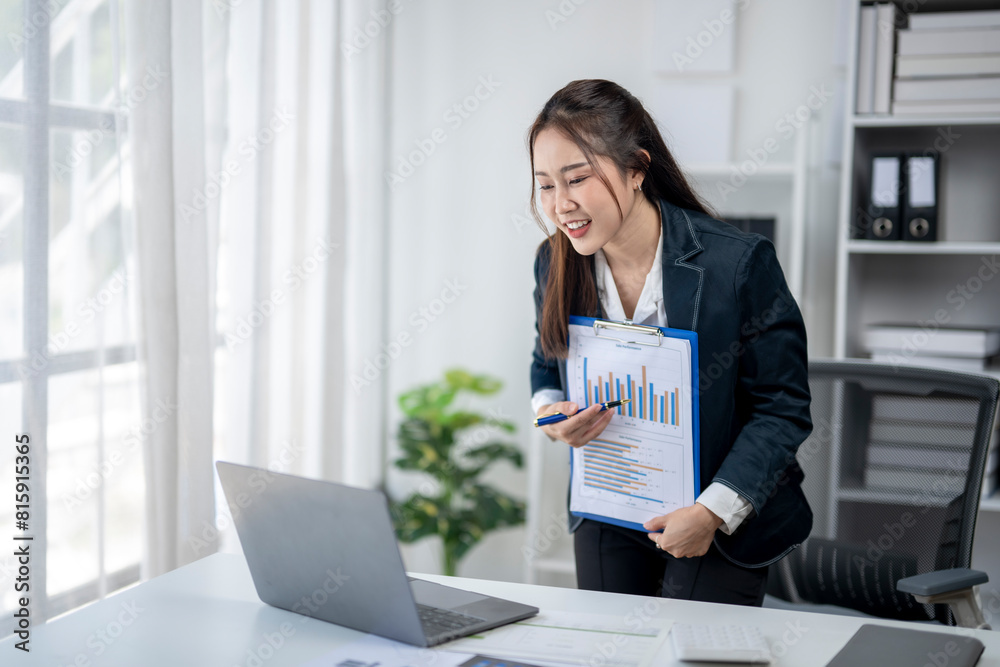 A woman in a business suit is standing in front of a laptop computer and holding a clipboard. She is excited or happy about something, possibly related to her work A woman in a business suit is standing in front of a laptop computer and holding a clipboard. She is excited or happy about something, possibly related to her work