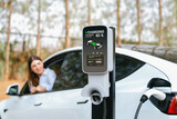 Focused EV charging station for electric vehicle's battery recharging on blurred background of young woman during her autumnal road trip travel. Eco friendly vacation with EV car during autumn. Exalt
