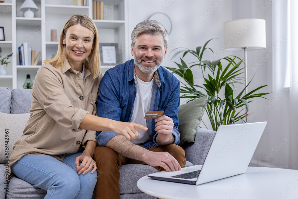 Joyful couple using a laptop and credit card for online shopping. They are sitting comfortably on a sofa in a cozy living room. Joyful couple using a laptop and credit card for online shopping. They are sitting comfortably on a sofa in a cozy living room.