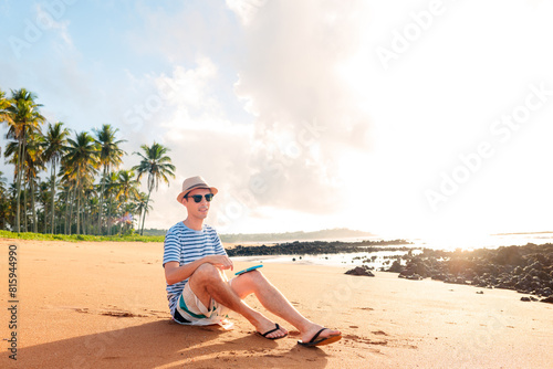 A man sitting on the sandy beach, relaxing. In the background, there's a paradisiacal scene with coconut trees and a sunrise