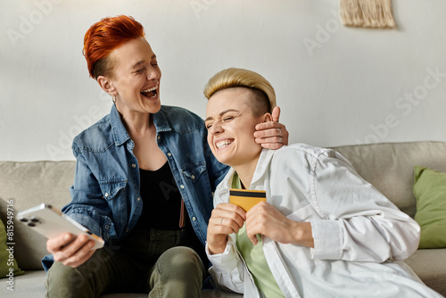 Two women with short hair sit on a couch, laughing while doing shopping online