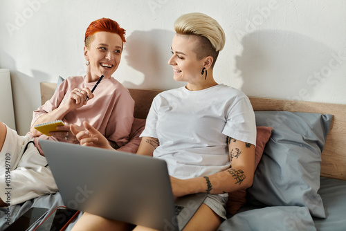 Two women with short hair sitting on a bed, engaged in using a laptop together.