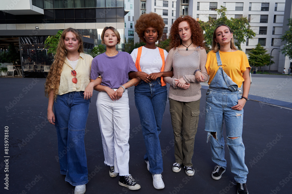 Obraz premium Multiethnic group of young women holding arm in arm looking at camera with seriousness. Concept of united female community, sorority, equality, feminism and empowerment.
